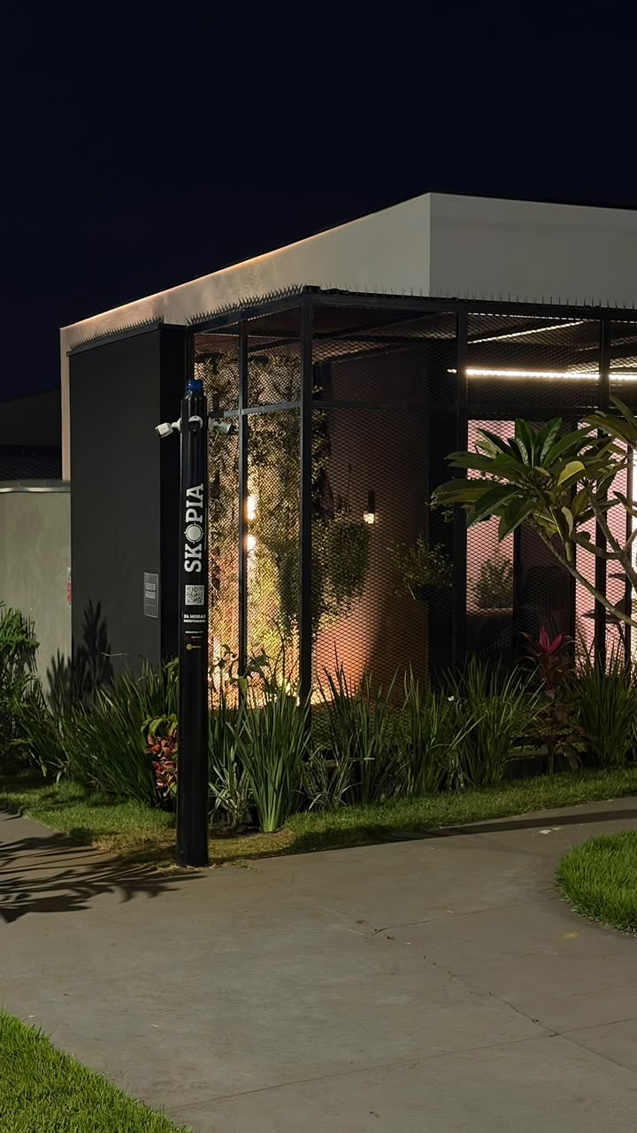 Fachada da QTI à noite com o totem de monitoramento SKOPIA na entrada, câmeras ativas e jardim iluminado